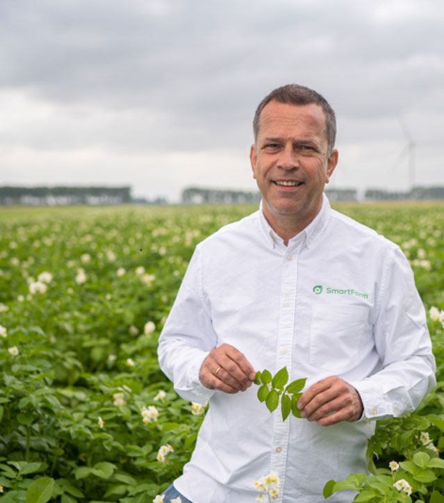 A man wearing a SmartFarm shirt stands in a field crop field while holding a twig